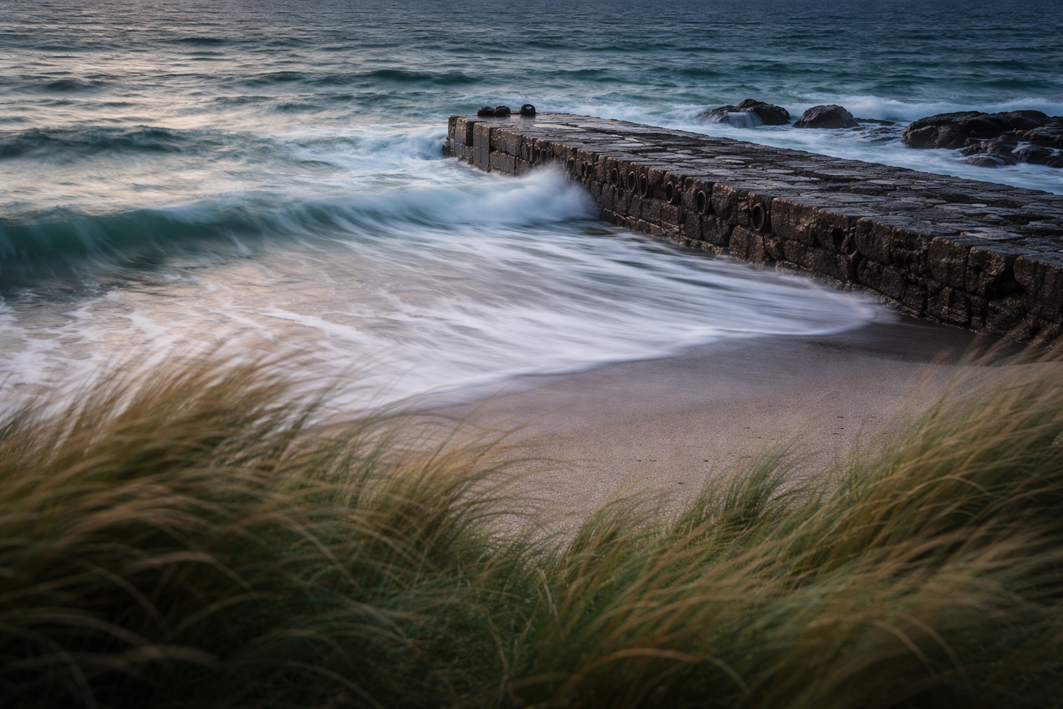 Long-exposure view of wind-blown coastal grasses and a streaked, foamy tide sliding over a weathered stone quay, with motion in the upper two thirds and a quiet, empty lower third.