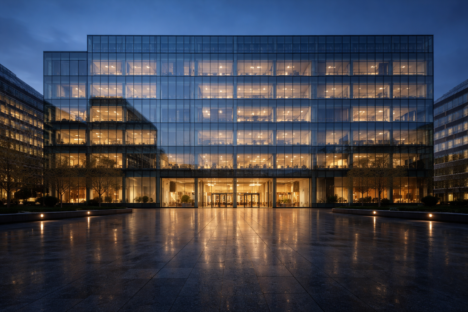 Wide evening view of a modern glass office building centered in the frame with warm interior lights visible through the curtain wall and an empty stone plaza in the lower foreground.