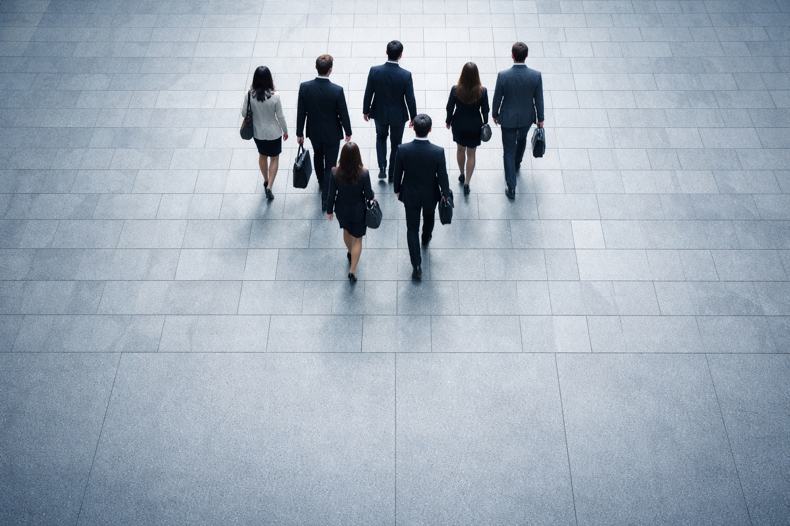 Top-down view of a few business-attired people crossing a paved city plaza, clustered in the upper two-thirds with empty paving in the lower third; faces are obscured from above.