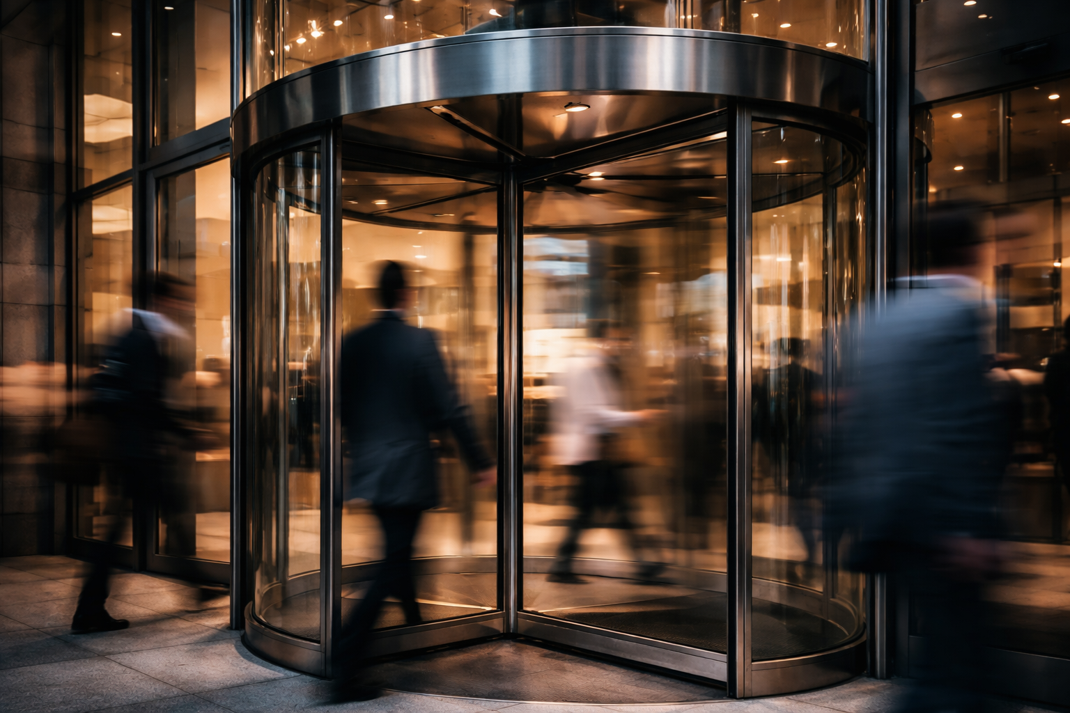 Dusk close-up of a revolving glass entrance: the stainless-steel hub and curved panels in sharp focus while anonymous commuters are motion-blurred around it, warm and cool reflections on the polished stone floor, lower third left uncluttered.