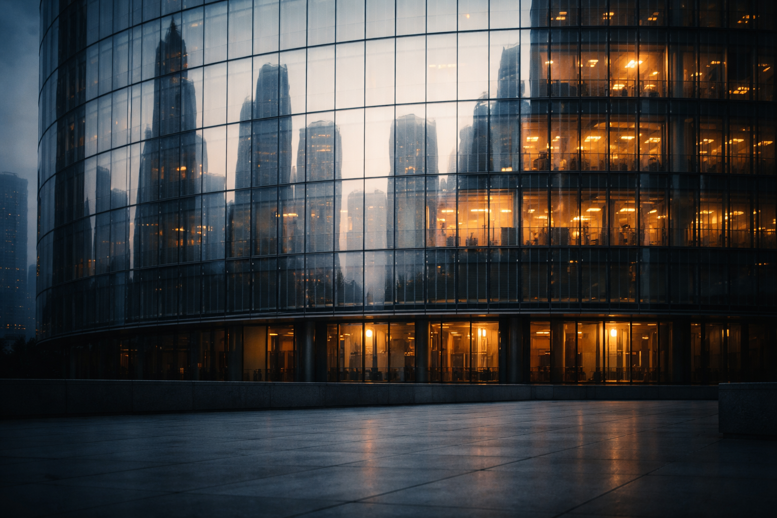 Curved glass facade of an anonymous office tower reflecting a blurred skyline and warm interior lights, with an empty stone plaza in shadow occupying the lower third.