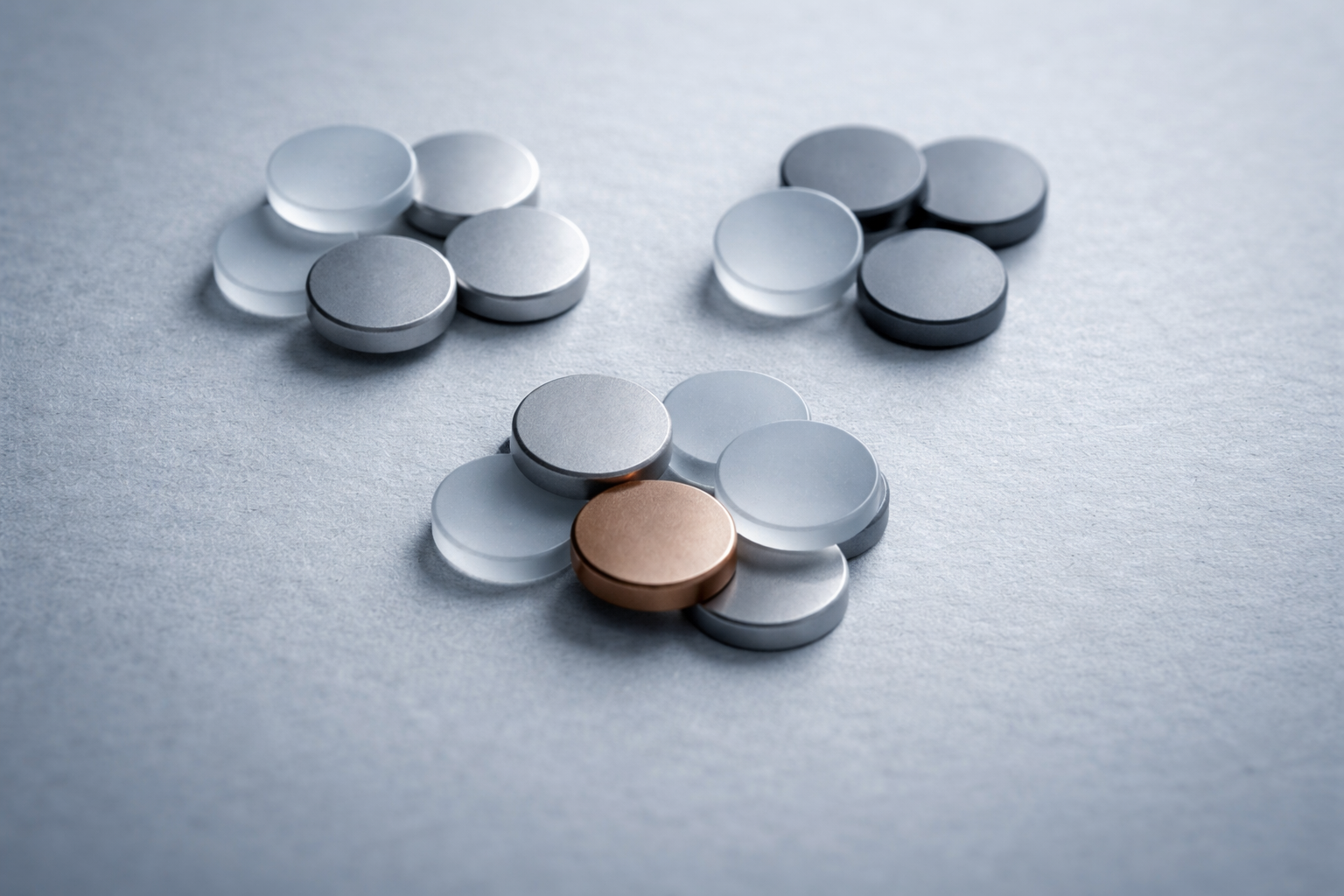 High-angle close-up of three small clusters of unbranded matte metal tokens and frosted translucent discs on a neutral textured tabletop, with soft cool light and an empty lower third.