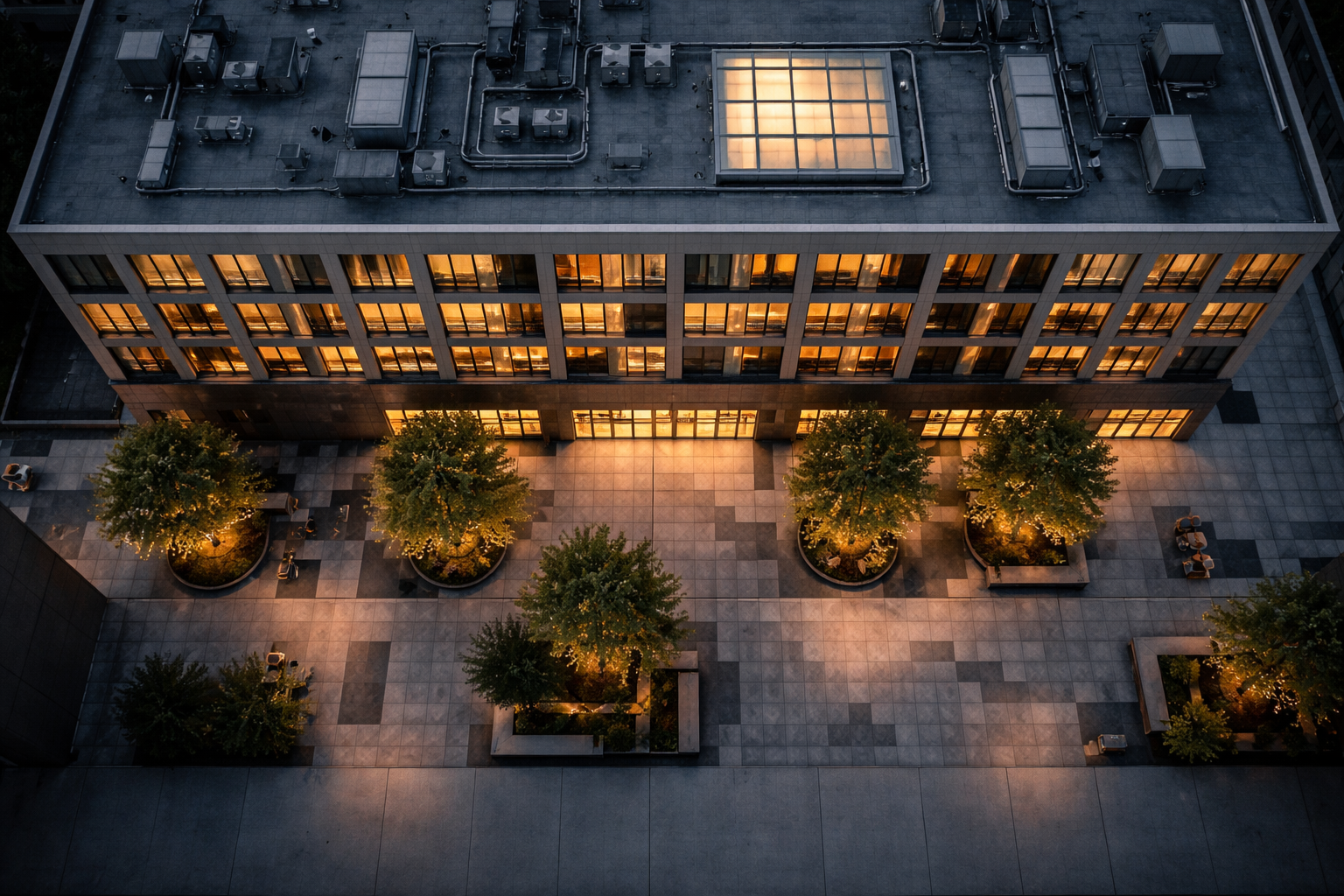 High-angle view from above of a mid-rise building rooftop and empty pedestrian plaza at dusk, with warm interior lights and cool blue twilight tones, geometric paving in the upper two thirds and a quiet uncluttered lower third.